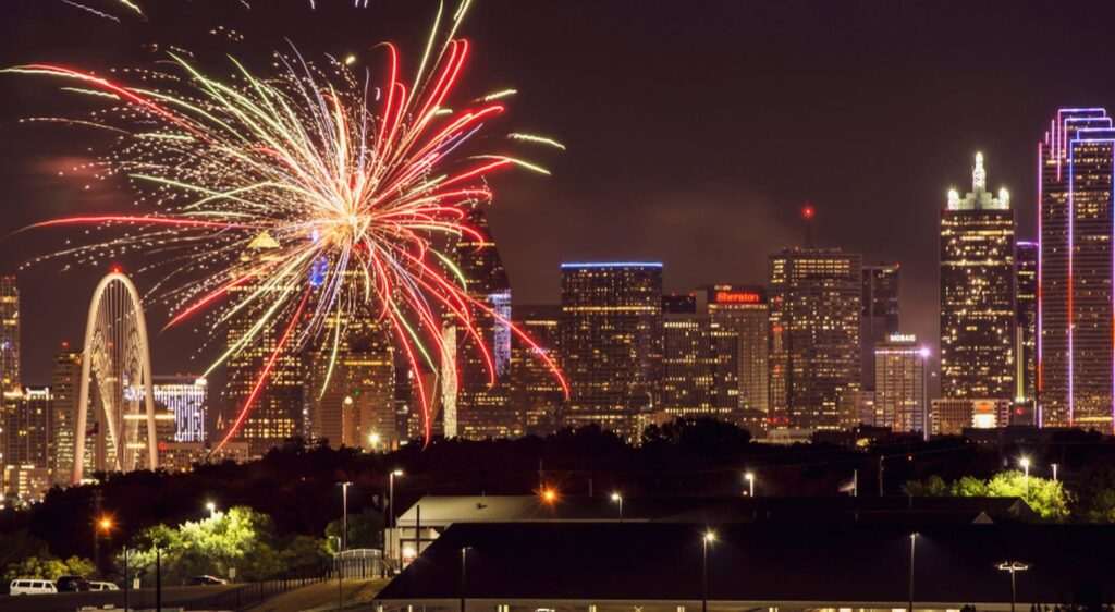 Colorful Fourth of July fireworks burst over the skyline at night, illuminating downtown skyscrapers and city landmarks.