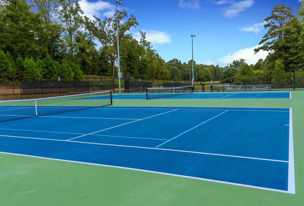 Well-maintained outdoor tennis courts with blue playing surfaces and green surrounds, featuring regulation court markings, tennis nets, perimeter fencing, and a wooded backdrop in a community recreation setting.