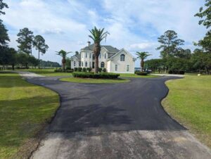 Newly paved circular asphalt driveway leading to a large custom home, featuring smooth black pavement, landscaped center island with palm trees, and expansive residential property grounds.