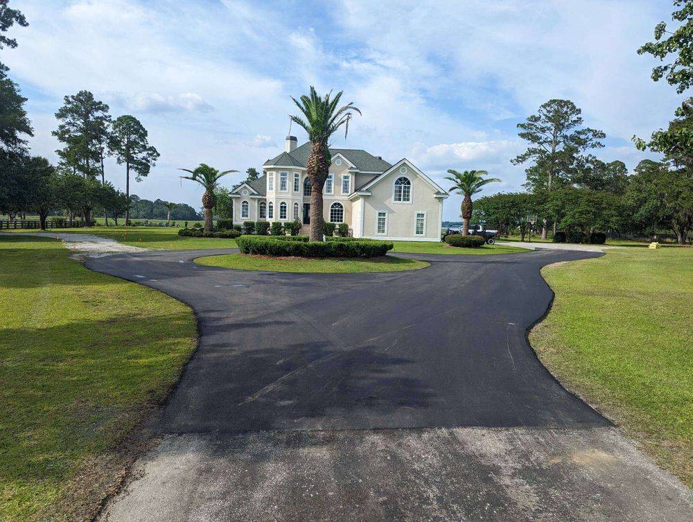 Newly paved circular asphalt driveway leading to a large custom home, featuring smooth black pavement, landscaped center island with palm trees, and expansive residential property grounds.